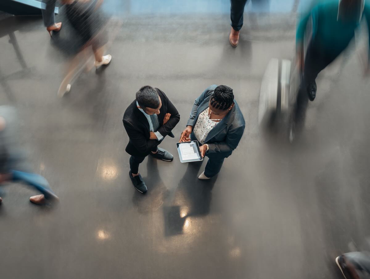 Two business people standing in the lobby of an office looking at a tablet while people are walking past in a blur 